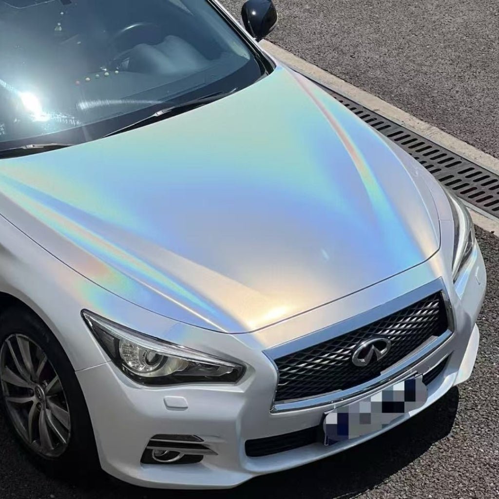 A white Infiniti sports a stunning iridescent hood and front with White Galaxy Glossy SelfRepair Nano-Coated PPF, reflecting pastel colors in the sunlight as it’s parked by a drainage grate.