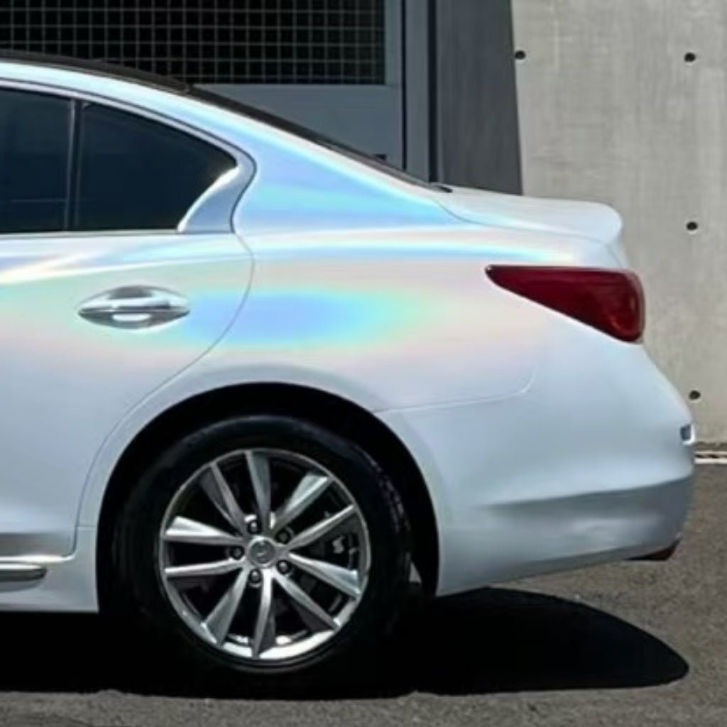 A white car shines with an iridescent rainbow reflection from White Galaxy Glossy SelfRepair Nano-Coated PPF, offering dokonalá ochrana karosérie; it’s parked by a concrete wall and metal gate, rear half in view.