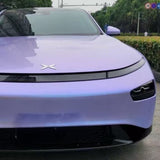 A close-up of the front of a sleek, silver-to-purple matte electric car with narrow headlights and a minimalist logo showcases Charm Silver to Purple Matte SelfRepair Nano-Coated PPF for added protection, parked on a city street with greenery and signs.