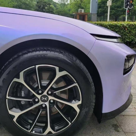 Close-up of the front left wheel and fender, featuring Charm Silver to Purple Matte SelfRepair Nano-Coated PPF on a modern car with black alloy rims, silver pattern, Brembo brakes, and greenery visible in the background.