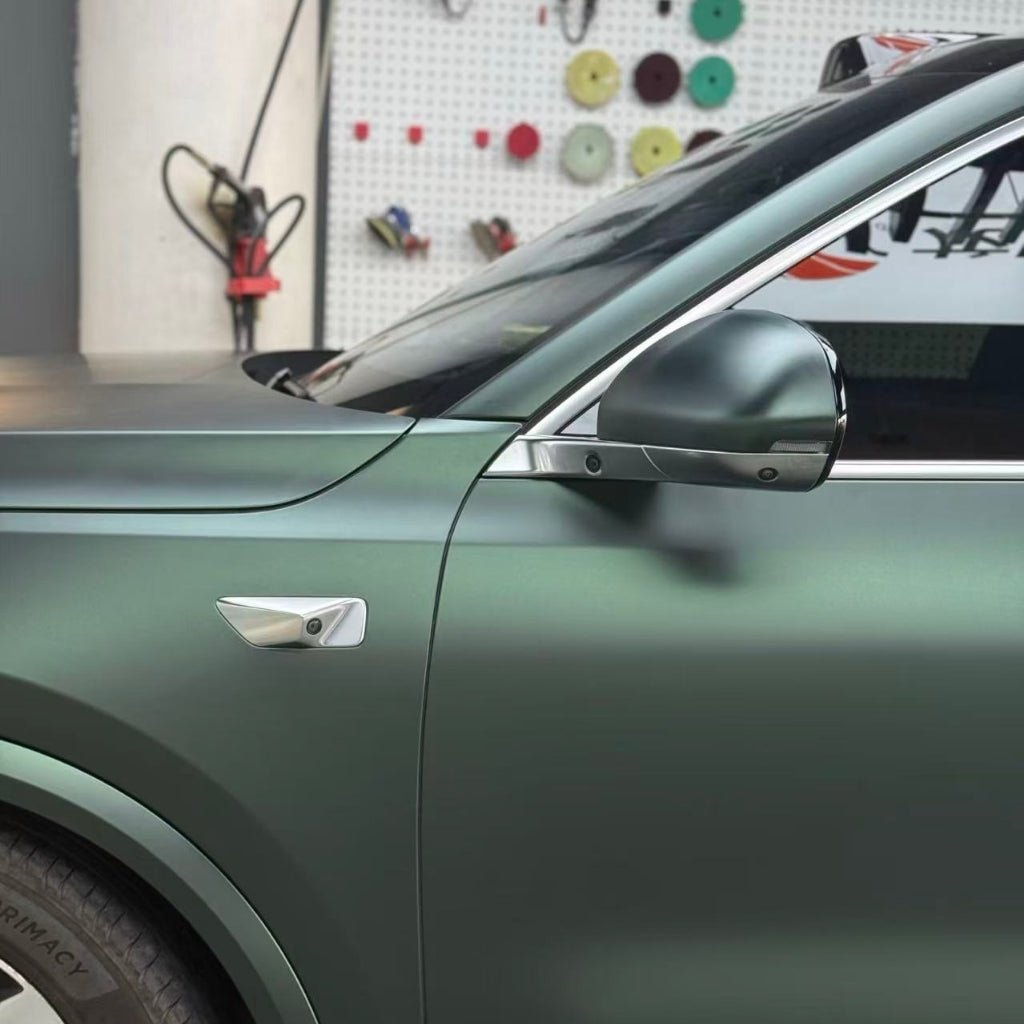 A close-up of a car’s front side wrapped in Dark Green Matte SelfRepair Nano-Coated PPF, highlighting the side mirror and part of the front wheel, with tools and supplies hanging on a white pegboard wall in the background.