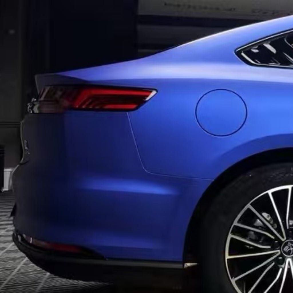 The image shows the rear side of a blue car wrapped in Diamond Blue Matte SelfRepair Nano-Coated PPF, highlighting the taillight, wheel, fuel cap, and back door under indoor lighting that emphasizes its sleek design and protective finish.