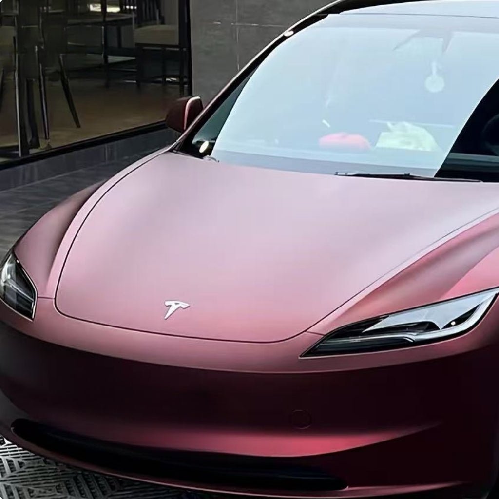 A Tesla with Dragon Blood Red Matte SelfRepair Nano-Coated PPF is parked outdoors by a building with glass windows, its front hood, headlights, and Tesla logo enhanced by the striking matte protective film.