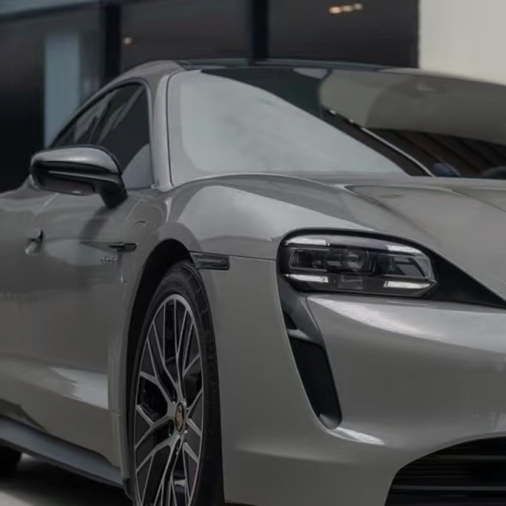 Close-up shot of the front left side of a sleek silver sports car with modern LED headlights and alloy wheels, featuring Porsche Gray Matte SelfRepair Nano-Coated PPF, parked in front of a contemporary building.