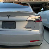 A silver Tesla with Dual Motor badge, protected by Nardo Gray Matte SelfRepair Nano-Coated PPF, is parked in a driveway next to a white car. Its rear license plate holder is empty, and there's a garage and house visible in the background.