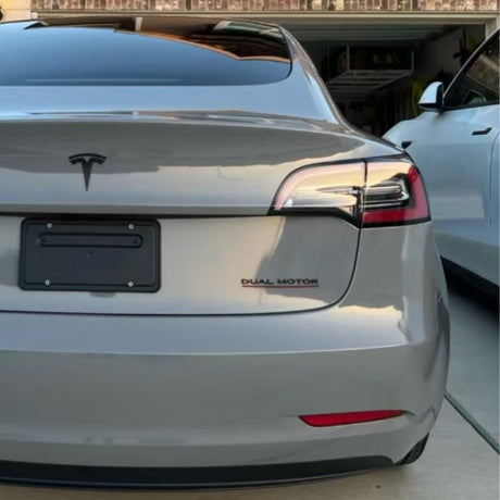 A silver Tesla with Dual Motor badge, protected by Nardo Gray Matte SelfRepair Nano-Coated PPF, is parked in a driveway next to a white car. Its rear license plate holder is empty, and there's a garage and house visible in the background.