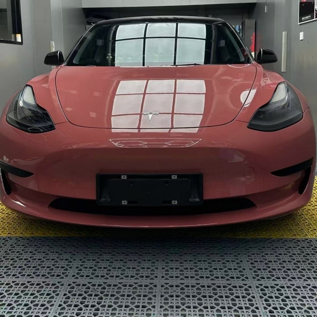A front view of a pink Tesla Model 3 in Morganite Red Glossy SelfRepair Nano-Coated PPF, parked indoors as ceiling grid windows reflect on its hood, highlighting advanced body protection and self-healing nano technology.