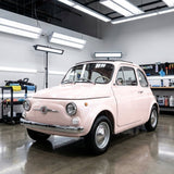 A vintage light pink Fiat 500, protected by Pearl Blossom Pink Glossy SelfRepair Nano-Coated Nexthor™ PPF, is parked in a modern garage surrounded by bright lights and detailing equipment.