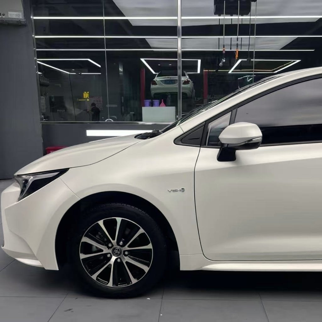 A car protected with Satin Liquid Metallic Bronze SelfRepair Nano-Coated PPF, featuring black alloy wheels, is parked indoors under ceiling lights. The front door, mirror, and hood are visible with a glass wall and another vehicle in the background.
