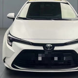 Front view of a white Toyota sedan indoors, showcasing its grille, headlights, and Toyota emblem. The car is protected with Satin Liquid Metallic Bronze SelfRepair Nano-Coated PPF. Neutral gray background.