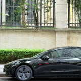 A sleek car protected by Shell Black Glossy SelfRepair Nano-Coated PPF is parked beside a stone wall with iron railings and greenery, showcasing its glossy finish on the front half—door, mirror, and wheel—in the photo.