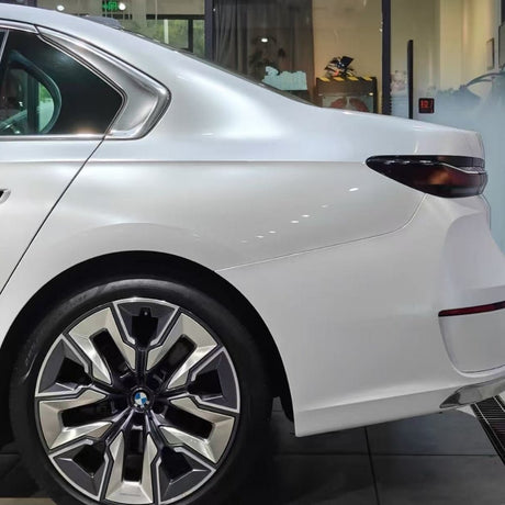 The image shows the rear of a white BMW sedan with Shell White Glossy SelfRepair Nano-Coated PPF, highlighting the back door, rear fender, and a large geometric alloy wheel. Glass walls and vivid reflections fill the background.