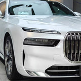 Close-up of a white luxury car with Shell White Glossy SelfRepair Nano-Coated PPF on the front left, highlighting the sleek headlight, chrome grille, and wheel. A paper sign is visible on the dashboard through the windshield.