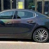 Side view of a black luxury sedan on the street, its paint protected by Stellar Black Glossy SelfRepair Nano-Coated PPF, featuring chrome trim and large alloy wheels. A building entrance appears in the background.