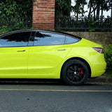 A bright yellow electric car with black wheels, protected by São Paulo Glossy SelfRepair Nano-Coated PPF, is parked beside a brick pillar and stone wall, with greenery and trees in the background.