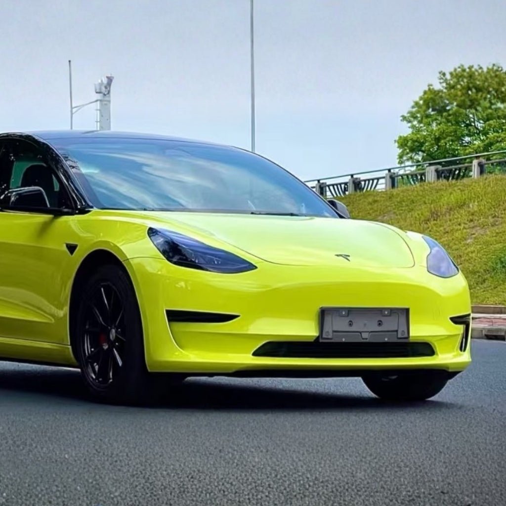 A Tesla Model 3 in bright yellow-green with black wheels is parked on a paved road, its vivid finish shielded by São Paulo Glossy SelfRepair Nano-Coated PPF. Green grass and a tree form a picturesque background.