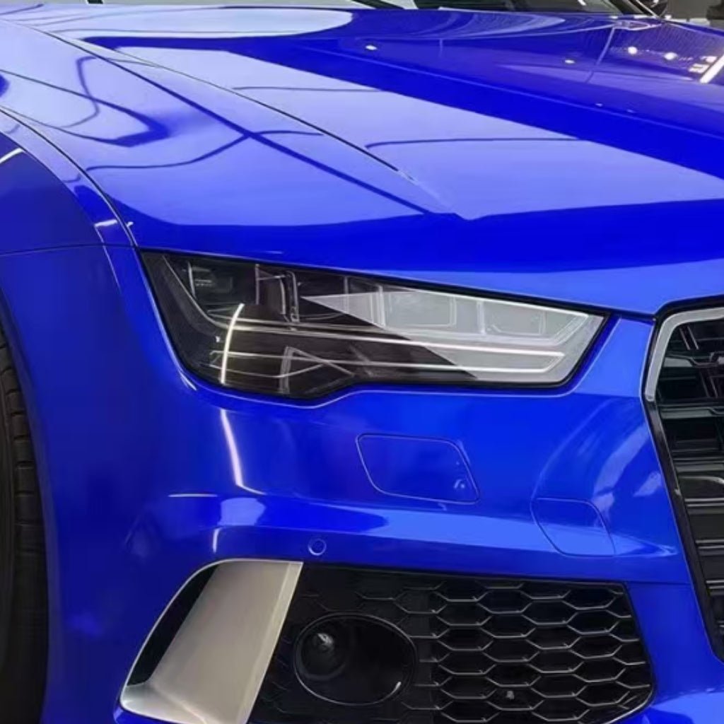 Close-up of the front left side of a shiny blue car with Sapphire Glossy SelfRepair Glossy Nano-Coated PPF, showing the headlight, hood, grille, and bumper reflecting lights on its self-healing nano-treated surface.