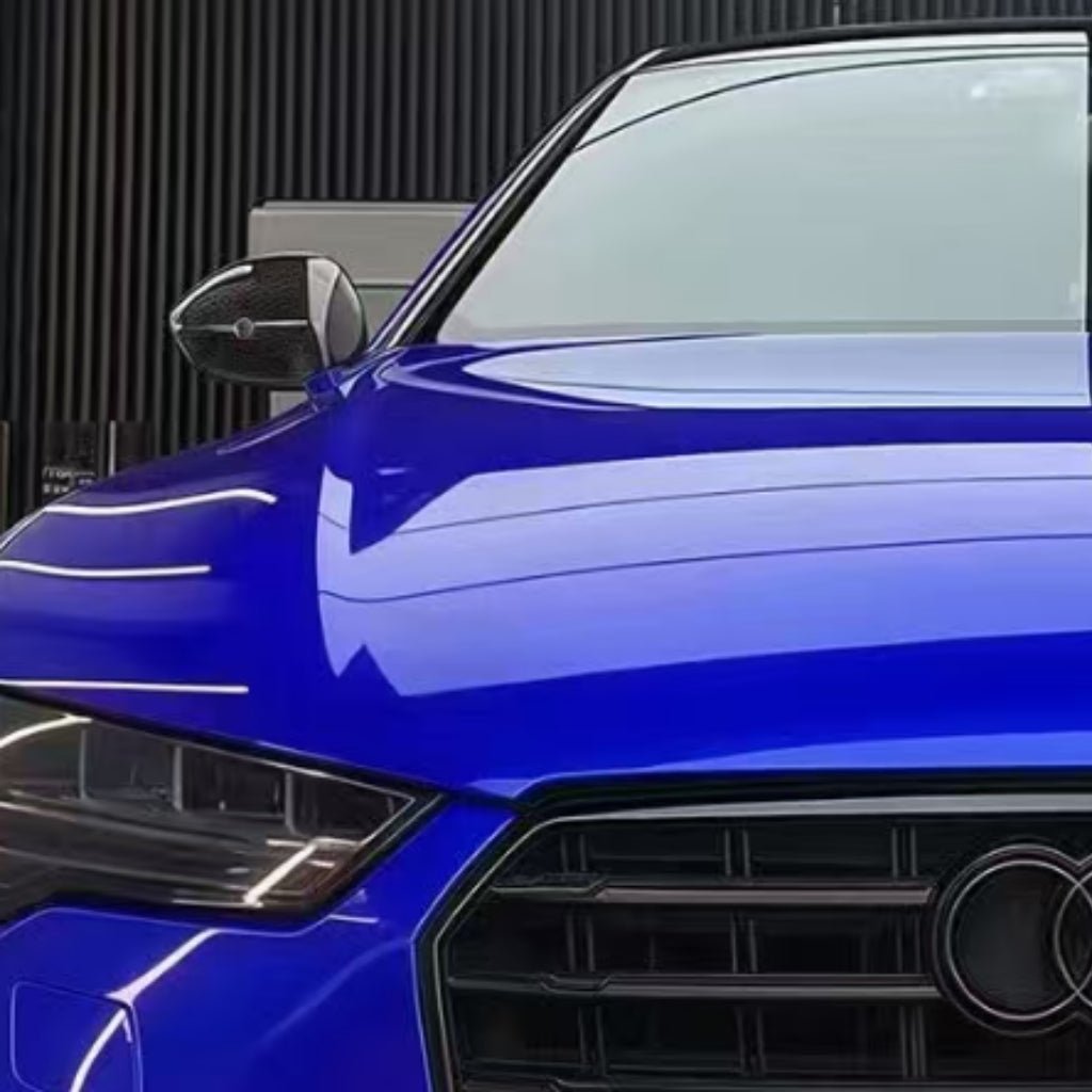 Close-up of a shiny blue car's front end, featuring the hood, headlight, grill, and side mirror protected by Sapphire Glossy SelfRepair Glossy Nano-Coated PPF, set against a dark striped background.