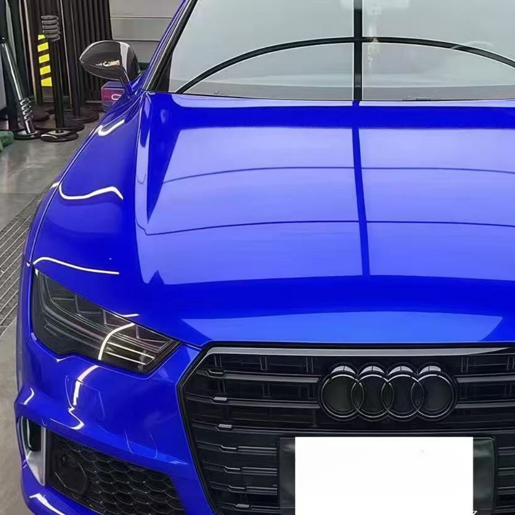A close-up of a blue Audi’s front shows the grille, headlights, and hood covered in Sapphire Glossy SelfRepair Glossy Nano-Coated PPF, protecting the iconic four-ring logo under bright indoor lights.