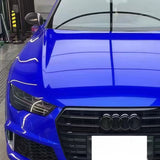 A close-up of a blue Audi’s front shows the grille, headlights, and hood covered in Sapphire Glossy SelfRepair Glossy Nano-Coated PPF, protecting the iconic four-ring logo under bright indoor lights.