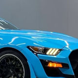Close-up of the front left side of a bright blue sports car protected by Sky Blue Glossy SelfRepair Nano-Coated PPF, showcasing a black wheel, triple-striped illuminated headlight, and amber turn signal on a plain gray background.