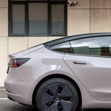 The rear of a light-colored Tesla Model 3 with Sky Mirror Pink Glossy SelfRepair Nano-Coated PPF, parked on the street; a modern beige building with a black-framed window appears in the background.