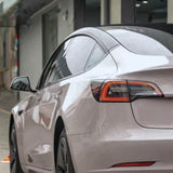 A rear side view of a light-colored Tesla Model 3 with Sky Mirror Pink Glossy SelfRepair Nano-Coated PPF, parked outdoors near a building, showcasing the car’s sleek design, taillight, and rear wheel.