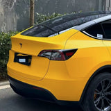 A yellow Tesla with tinted windows, protected by Sunshine Yellow Glossy SelfRepair Nano-Coated PPF, is parked on the street near green bushes and a gray wall. The image highlights the car's rear and side.
