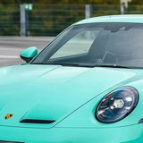 A close-up shows the front of a turquoise Porsche featuring Tiffany Blue Glossy SelfRepair Nano-Coated PPF on its hood, left headlight, windshield, and side mirror. The car is parked outdoors with a blurred fence and greenery in the background.