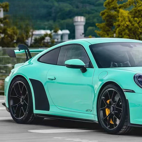 A Porsche 911 GT3 with Tiffany Blue Glossy SelfRepair Nano-Coated PPF is parked outdoors by greenery and a fence, featuring black accents, a large rear wing, black alloy wheels, and yellow brake calipers for dokonalá ochrana laku vozidla.