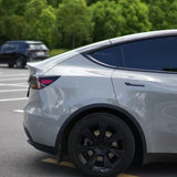 A silver car, partially shown from the side and protected with Volcano Gray Glossy SelfRepair Nano-Coated PPF, is parked in a sunny parking lot with surrounding trees. In the background, a black car adds to the scene's calm atmosphere.