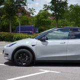 A silver electric car with black wheels, protected by Volcano Gray Glossy SelfRepair Nano-Coated PPF, is parked in a lot near trees and a blue van in the background.