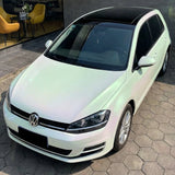 A white Volkswagen Golf hatchback with a black panoramic roof, wrapped in White Diamond Green Glossy SelfRepair Nano-Coated PPF, is parked on hexagonal pavement near a modern building with yellow chairs and a glass wall.