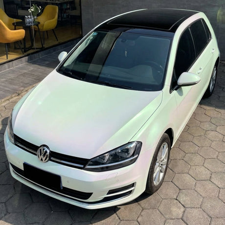 A white Volkswagen Golf hatchback with a black panoramic roof, wrapped in White Diamond Green Glossy SelfRepair Nano-Coated PPF, is parked on hexagonal pavement near a modern building with yellow chairs and a glass wall.