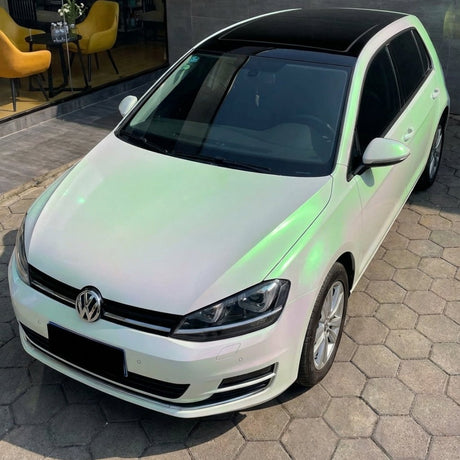 A white Volkswagen Golf hatchback with a black roof, protected by White Diamond Pine Green Glossy SelfRepair Nano-Coated PPF, is parked on a stone-tiled driveway near a building with large windows and yellow chairs inside.