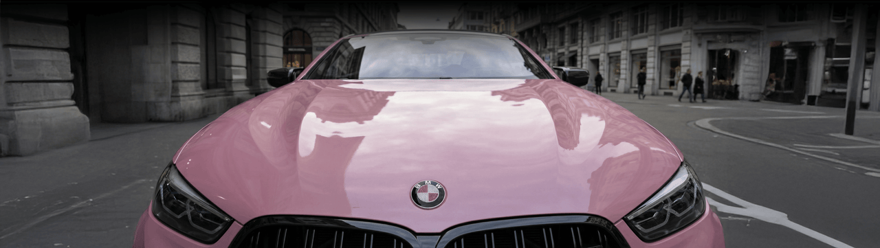 A shiny pink BMW car is parked on a city street, reflecting buildings on its hood. The photo is taken from the front, showing the bold BMW logo and urban surroundings.