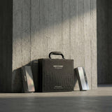 A black textured case labeled Nexthor PPF Sample Kit (All Colors) stands upright against a concrete pillar, with two rectangular metallic LED devices on the floor on either side. Sunlight casts soft shadows across the scene.