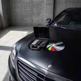 The Nexthor PPF Sample Kit (All Colors) is displayed on the matte black hood of a luxury car parked in a concrete garage, alongside a sleek black briefcase.
