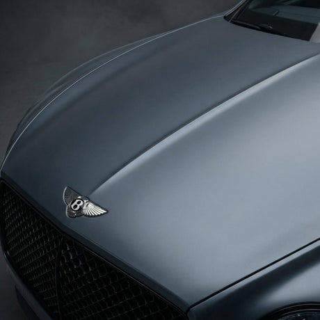 Close-up of the hood and front grille of a Bentley in Satin Titanium Ice, featuring the iconic winged B emblem and protected by Satin Titanium Ice SelfRepair Nano-Coated PPF, set against a dark, blurred background.